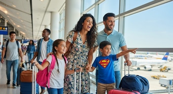 Una familia latina sonriente, con padre, madre, una niña y un niño, camina por un aeropuerto internacional con maletas, mirando por una ventana grande.