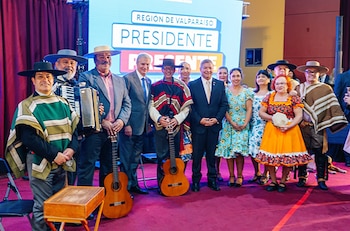 Un grupo de personas sonríe, algunas con vestimenta folclórica chilena y otras en traje, con guitarras, ante una pantalla que dice "Región de Valparaíso"
