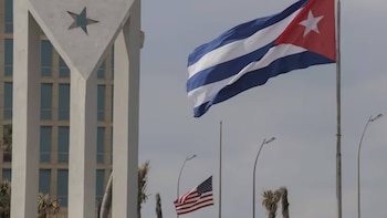 Bandera de Cuba ondeando en primer plano con un edificio de fondo que tiene una estrella. Debajo, una bandera de Estados Unidos también ondea