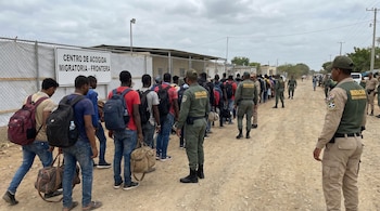 Una larga fila de hombres con mochilas y bolsos espera en un camino de tierra. Agentes de Migración de la República Dominicana los supervisan junto a una valla con un cartel que indica un centro de acogida.