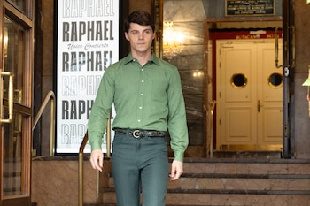Un hombre con camisa verde y pantalón oscuro camina cerca de un cartel publicitario con la palabra Raphael