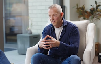 Gustav Söderström, un hombre de mediana edad con cabello canoso, viste camisa azul y jeans, sentado en un sillón blanco con las manos entrelazadas