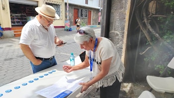 Hombre con sombrero y mujer con cabello canoso firman papeles en mesa blanca. Hay insignias azules, botella de agua. Al fondo, calle con edificios