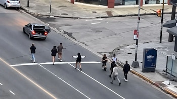 Varias personas cruzan una calle asfaltada cerca de una esquina mientras un automóvil gris permanece detenido con el baúl abierto