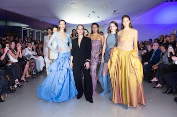 Un grupo de modelos con vestidos de gala posa en una pasarela junto a una persona en traje negro frente a los asistentes