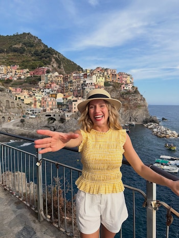 Mujer sonriente con sombrero de paja, blusa amarilla y shorts blancos extiende la mano. De fondo, pueblo costero con casas coloridas en acantilado y mar con barcos