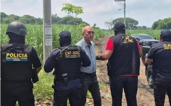 Cuatro agentes uniformados, dos con pasamontañas, y un hombre con camisa a cuadros en un camino de tierra. Un vehículo oscuro al fondo