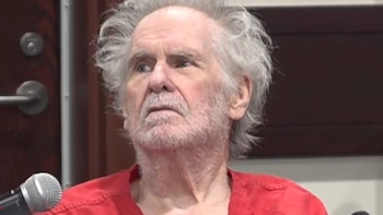 Hombre de edad avanzada con barba y cabello canoso, vestido con uniforme carcelario rojo frente a un micrófono
