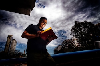 Hombre sonriente con gafas y polo oscuro lee un libro iluminado, de frente, bajo un cielo nublado y vibrante, con edificios y árboles urbanos al fondo