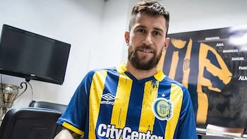 Jonatan Gómez, hombre barbudo, sonríe llevando camiseta de fútbol azul y amarilla. Fondo desenfocado con TV, trofeo y póster