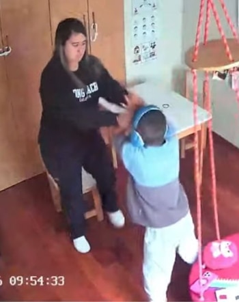 Una mujer y un niño en una habitación con piso de madera. La mujer viste ropa oscura. El niño usa ropa clara y auriculares azules. Se ve una mesa y un columpio