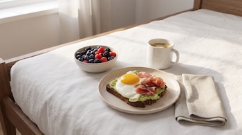 Plato con tostada de aguacate, huevo frito y jamón, cuenco con frutos rojos, taza de café y servilleta colocados sobre la ropa de cama.