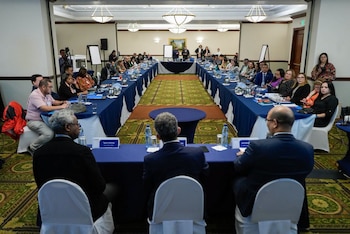 Sala de conferencias, mesa larga en forma de U, varias personas sentadas, botellas de agua, vasos, documentos, dos hombres de pie al frente