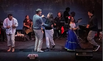 Varias personas cantan, tocan instrumentos y bailan sobre un escenario, entre ellas una mujer con falda azul y una pareja en el centro