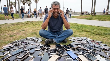 Hombre latinoamericano sonriente sentado en el pasto, con dos celulares en las orejas, rodeado de una gran pila de móviles. En el fondo se ven personas y palmeras.
