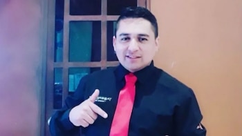 Hombre con camisa negra y corbata roja señala con el dedo hacia su pecho frente a una ventana y una pared de color marrón