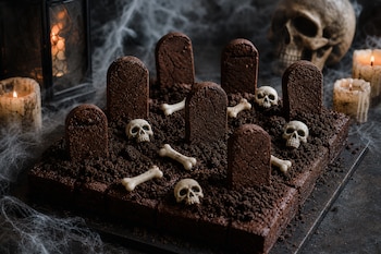 Brownies de chocolate cubiertos con migajas, figuras de lápidas, calaveras y huesos pequeños, rodeados por velas encendidas y telarañas.