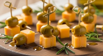 Aceitunas verdes y cubos de queso amarillo ensartados en palillos, ramitas de romero fresco y gotas de aceite sobre una tabla de madera oscura.