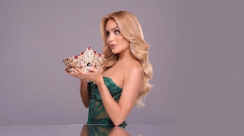 Una mujer con cabello rubio ondulado y vestido verde sostiene una corona dorada con piedras rojas y blancas.