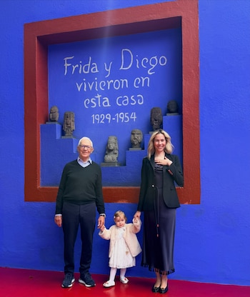 Un hombre, una niña y una mujer de pie frente a una pared azul con una inscripción sobre Frida y Diego y varias figuras de piedra