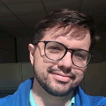 Primer plano de Joshua LeBlanc, un hombre caucásico de pelo oscuro y barba corta, usando gafas de montura negra y una chaqueta azul, sonriendo