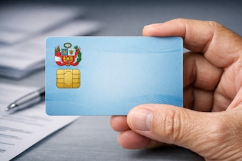 Primer plano de una mano de adulto mayor que sostiene un DNI electrónico peruano azul celeste con chip dorado y escudo. Mesa con papeles y bolígrafo.