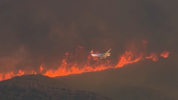 Un avión vuela cerca de una colina envuelta en humo denso y llamas de color naranja