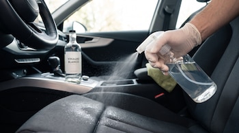 Mano con guante de látex sostiene atomizador rociando líquido sobre asiento de automóvil; botella de vinagre blanco en consola central, volante a la izquierda.