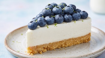 Porción de pastel con base de galleta, relleno blanco y cobertura de arándanos con gotas de agua sobre un plato.