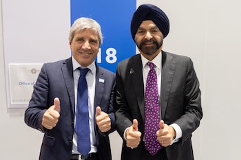 Luis Caputo y Ajay Banga visten traje formal y hacen el gesto de pulgares arriba frente a una pared blanca con un panel azul