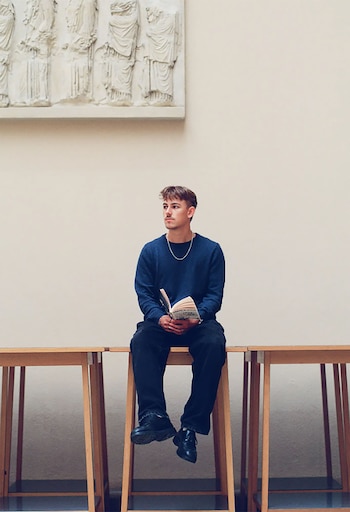 Nelio Biedermann, un joven de pelo castaño, está sentado en un banco de madera, sosteniendo un libro abierto. Detrás, una pared lisa con un relieve blanco