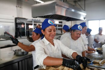 Una mujer sonriente con gorro y delantal blanco con un logo de comida, sostiene una bandeja negra con tres platos de comida. Al fondo, personas y pancartas