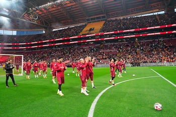 Vista aérea de jugadores del Galatasaray con uniforme granate aplaudiendo en el campo de un estadio lleno de aficionados. Las gradas son rojas y amarillas, y hay letreros de "RAMS PARK"