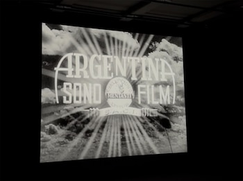 Proyección en blanco y negro del logo de Argentina Sono Film, con texto blanco 'Argentina Sono Film' sobre un fondo de nubes y rayos de luz