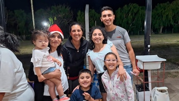 Siete personas, entre adultos y niños, posan de pie en un espacio exterior de noche; una de ellas lleva un gorro navideño y carga a una bebé