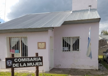 Edificio rosado de techo de chapa con ventanas enrejadas, un cartel con el texto Comisaría de la Mujer y una bandera argentina
