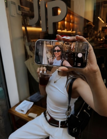 Una mujer sosteniendo un smartphone toma una selfie de sí misma sonriendo con gafas de sol y una bebida, reflejada en la pantalla, frente a un local con el letrero 'UP'.