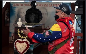 Un hombre con gorra y chaqueta colorida habla frente a un busto oscuro, un arreglo floral en forma de corazón y un cartel