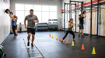 Personas en un gimnasio realizan ejercicios con una escalera de agilidad en el suelo, conos de colores y balones medicinales contra la pared.