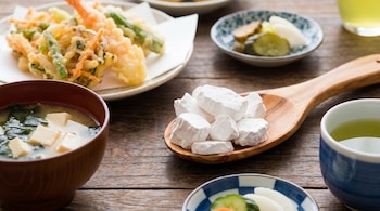 Una cuchara de madera sostiene bloques blancos de kuzu sobre una mesa rústica. Rodean la cuchara un plato de tempura, una sopa miso y cuencos de encurtidos.
