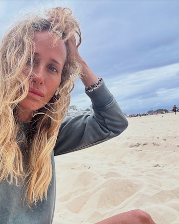 Una mujer con buzo gris y cabello rubio sentada en una playa de arena clara, bajo un cielo nublado y con personas a la distancia
