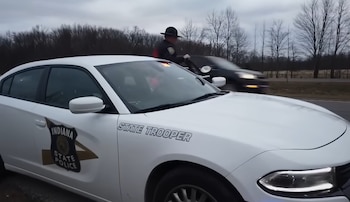 Oficial de policía de Indiana con sombrero de ala ancha junto a coche patrulla blanco en la orilla de una carretera, con un coche oscuro pasando velozmente