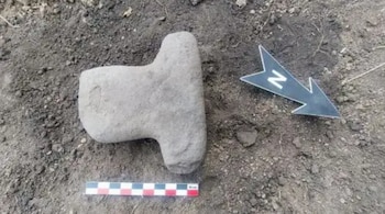 Un objeto tallado de piedra sobre la tierra junto a una escala métrica y una flecha negra que señala el norte