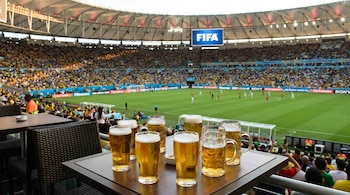 Varios vasos de cerveza sobre una mesa en un palco con vistas a un estadio de fútbol lleno de aficionados. En el fondo, una pantalla muestra el logo de FIFA.