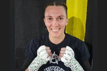 Mujer sonriendo con trenzas y camiseta oscura. Sus manos están vendadas con blanco y tienen logos verdes de WBC. Fondo negro y amarillo