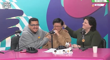 Tres personas, dos mujeres y un hombre, sentadas en una mesa rosa con tres micrófonos. Una mujer se cubre el rostro con la mano. Hay varias figuras y objetos sobre la mesa