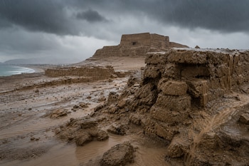 Estructuras de adobe erosionadas por corrientes de lodo durante la lluvia, con una edificación antigua y el mar al fondo.