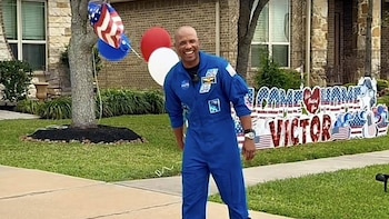 Un hombre calvo sonriente con un traje de vuelo azul de la NASA está en un jardín delantero con globos y un letrero de "Bienvenido a Casa Victor" con diseños de la bandera de EE. UU