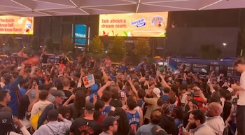 Vista de una gran concentración de personas en una plaza de la ciudad por la noche, con carteles digitales luminosos de Snapple y los Knicks, y un autobús de la ciudad
