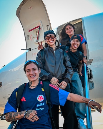 Cuatro mujeres con la indumentaria de la selección colombiana de fútbol posan en la puerta y escaleras de un avión